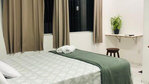 Une chambre à coucher avec un lit, des rideaux, une fenêtre, un bureau avec une plante et un petit tabouret.