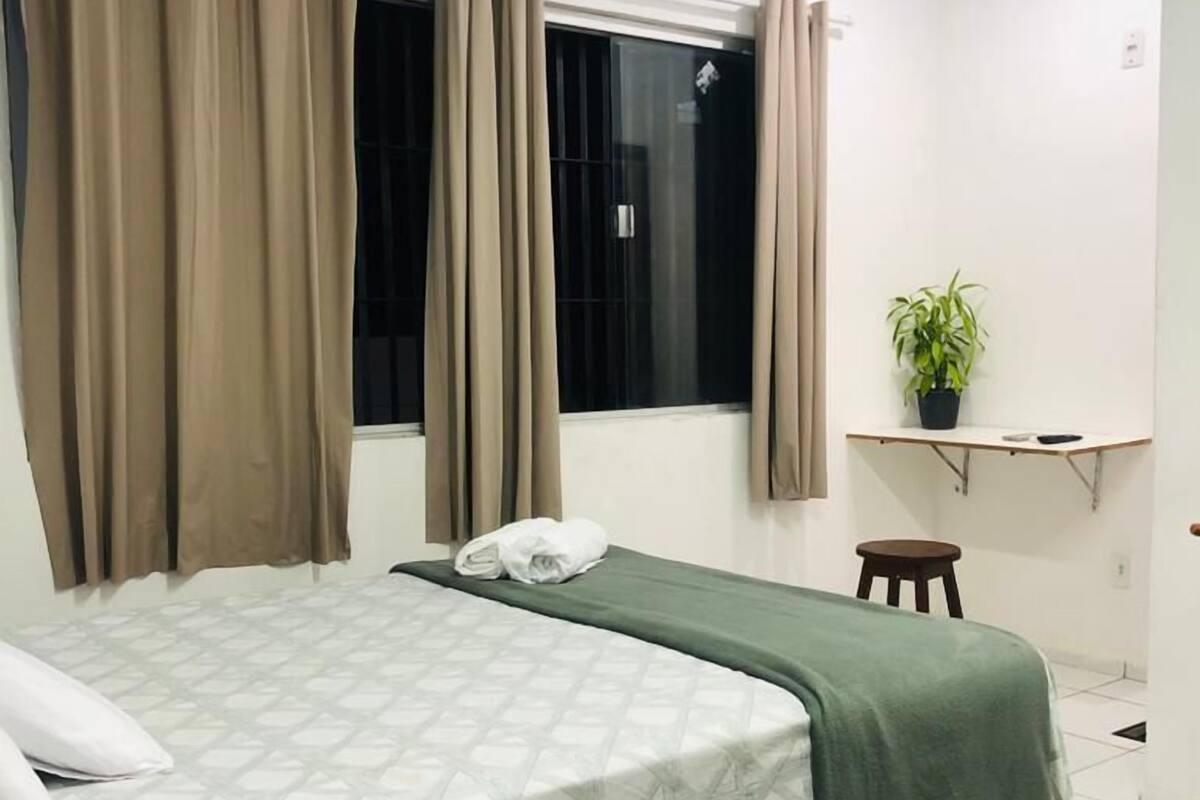 Un dormitorio con cama, cortinas, una ventana, un escritorio con una planta y un taburete pequeño.