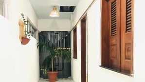 Un couloir étroit au sol carrelé de blanc, une plante en pot, un luminaire suspendu et une porte en bois avec des volets.