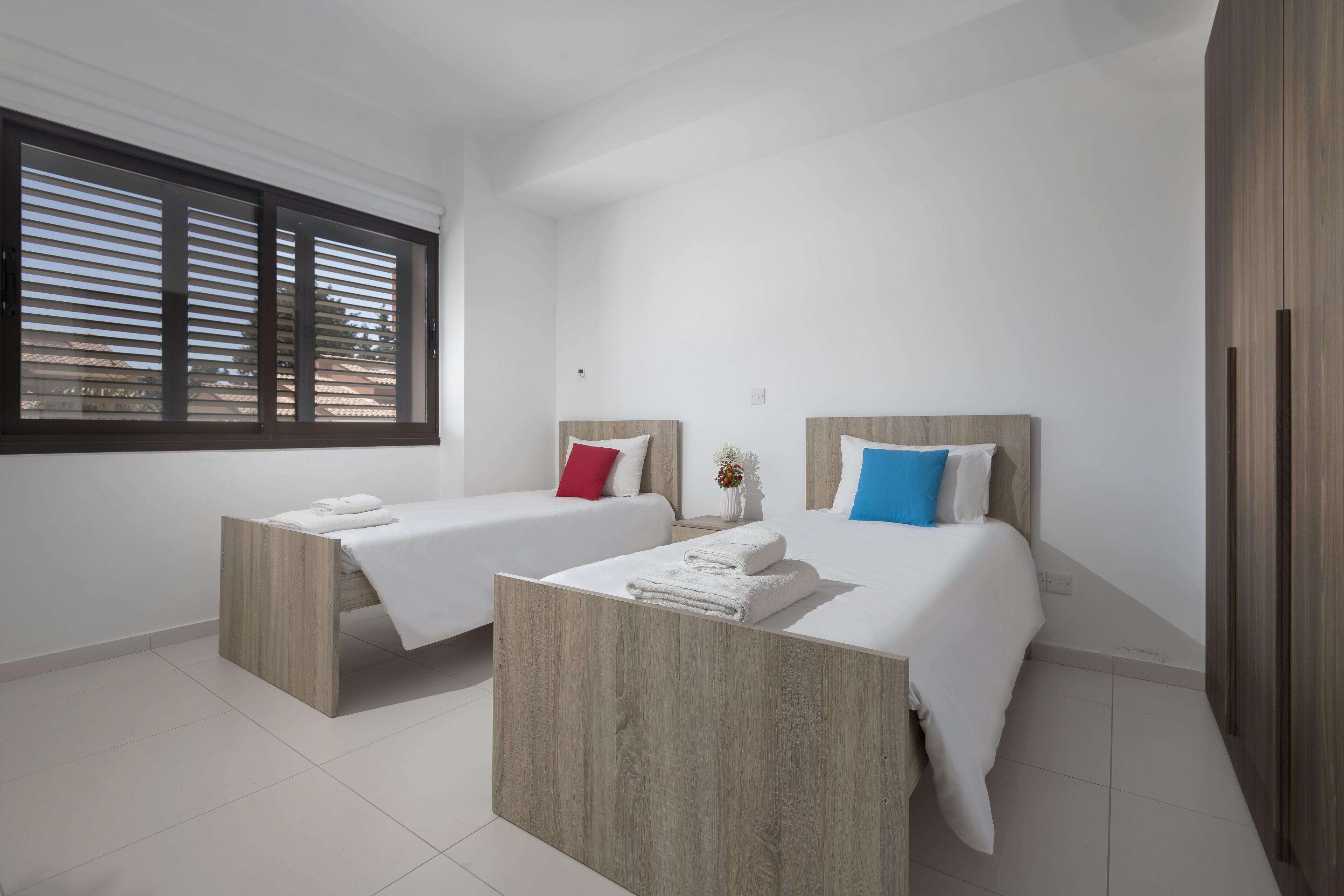 A modern bedroom with two beds, each with a wooden headboard, white bedding, and a small bedside table with a vase.