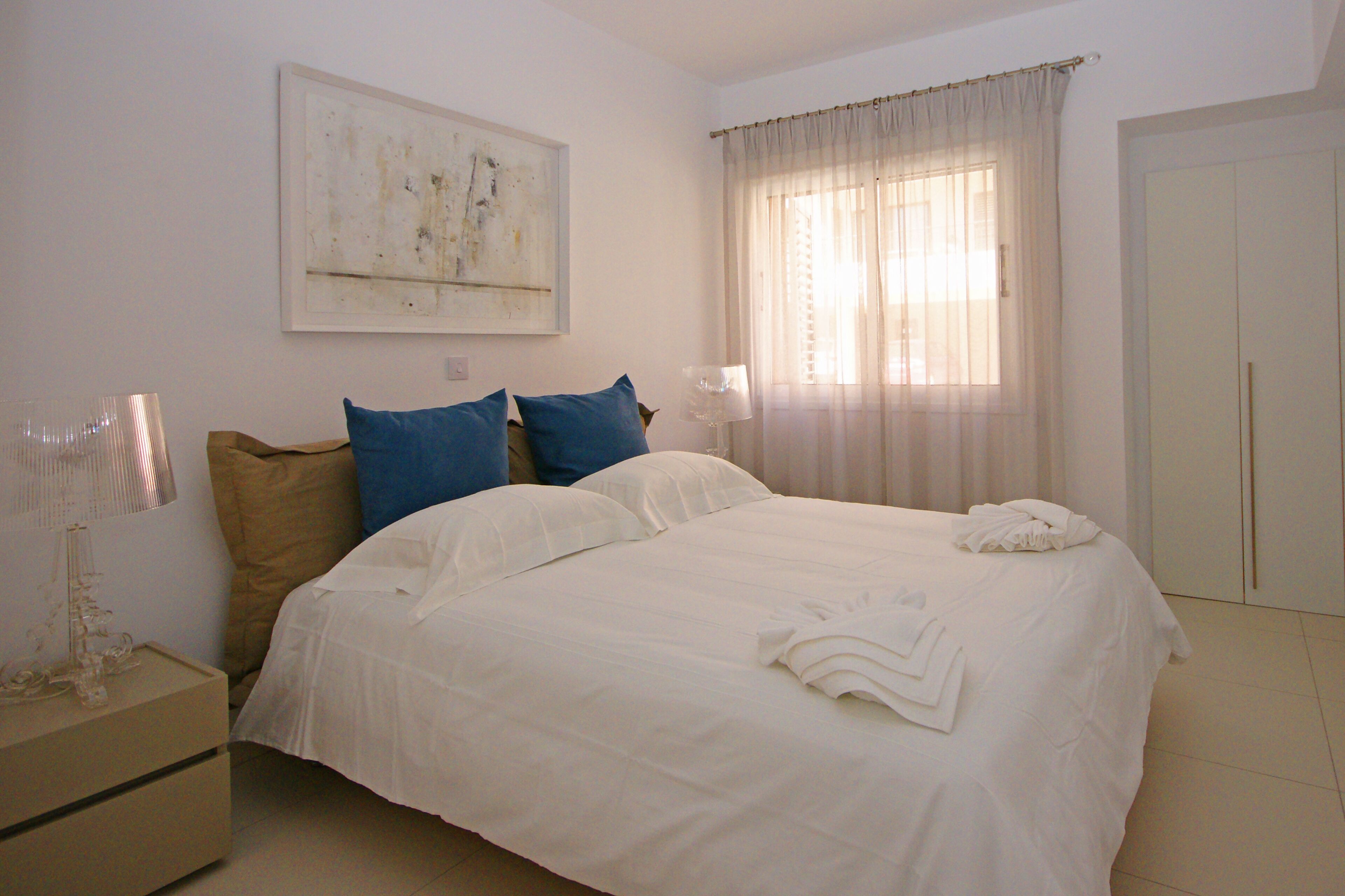Una cama bien hecha con ropa de cama blanca y azul, una mesita de noche con una lámpara y un cuadro en la pared.