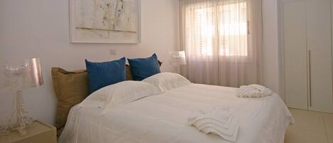 Una cama bien hecha con ropa de cama blanca y azul, una mesita de noche con una lámpara y un cuadro en la pared.