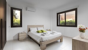 A modern bedroom with a wooden bed, white bedding, a bedside table, and a nightstand. There are two windows with black frames, one on each wall, and a small window air conditioning unit on the wall. - Resitour - Aphrodite Gardens (Paphos)