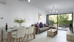 A modern living room with a dining area, a sofa with cushions, a coffee table, and a TV. - Resitour - Aphrodite Gardens (Paphos)