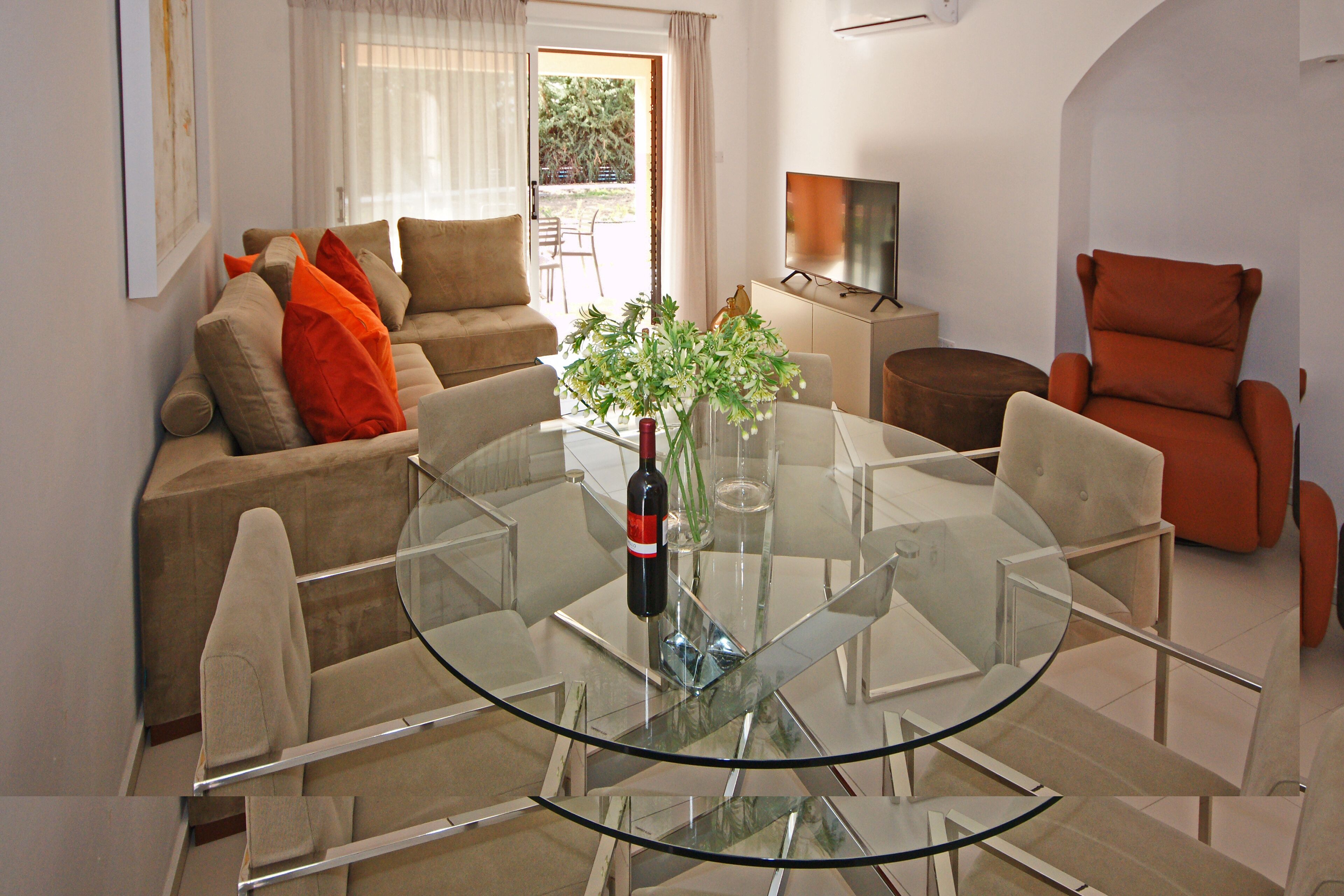 A modern living room with a glass-top dining table, beige sofas, and a red armchair.
