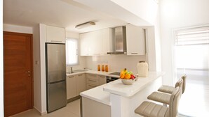 A modern kitchen with white cabinets, a stainless steel refrigerator, a fruit bowl on the counter, and a window with blinds. - Resitour - Aphrodite Gardens (Paphos)