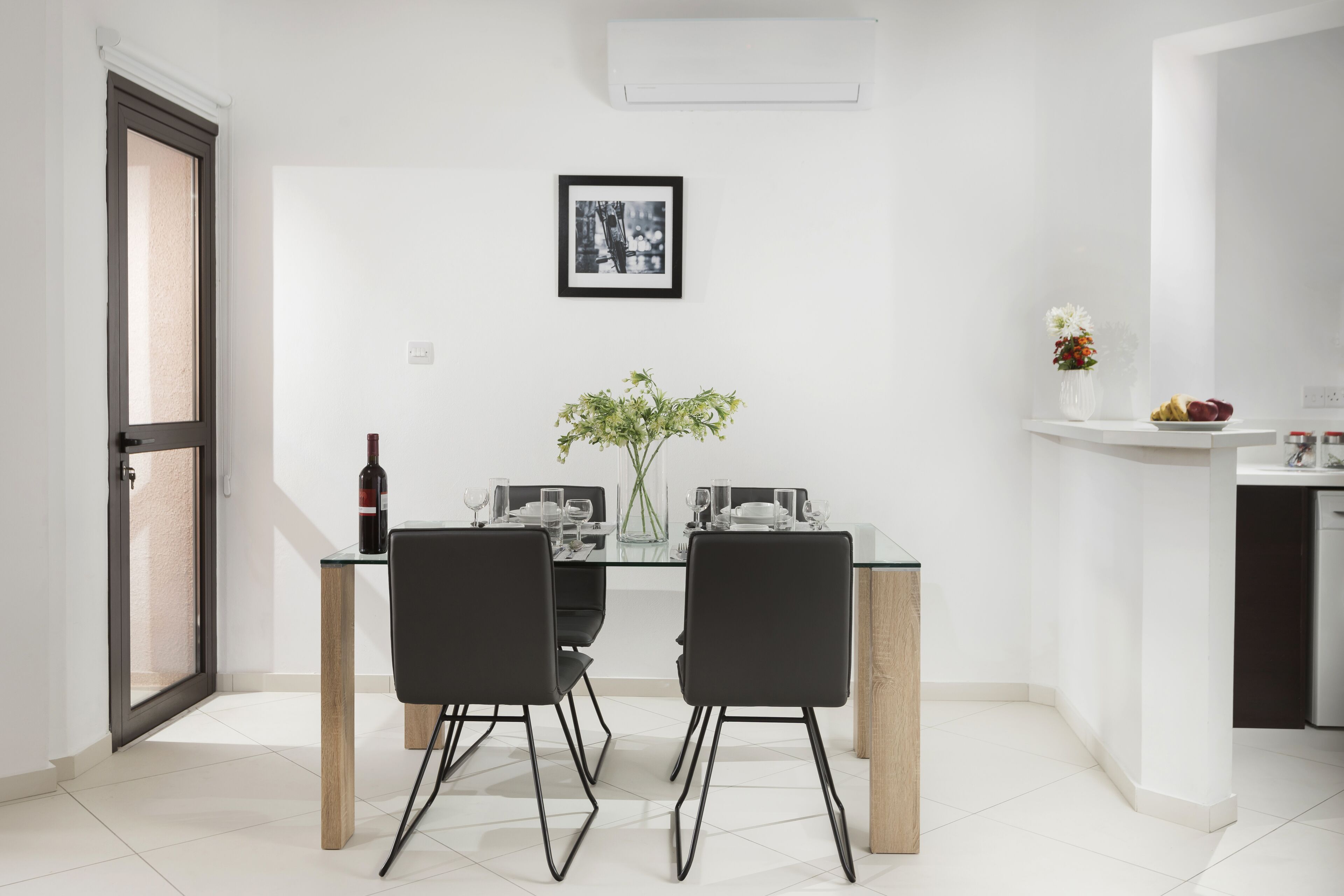 Un espace repas moderne avec une table en verre, deux chaises noires, un vase de fleurs et un tableau encadré au mur.