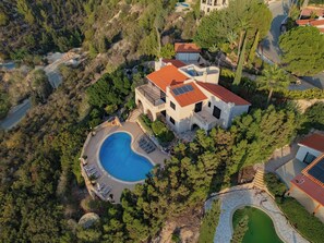 An aerial view of a villa with a swimming pool, surrounded by dense trees and a hilly landscape.