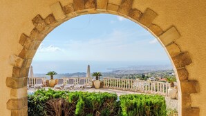 An arched stone structure with a view of a coastal landscape, including a city in the distance and a garden with chairs and potted plants. - Resitour - Kamares Village (Tala)
