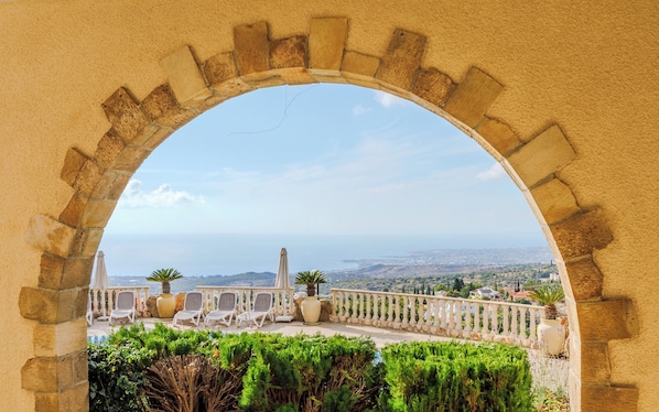 An arched stone structure with a view of a coastal landscape, including a city in the distance and a garden with chairs and potted plants.