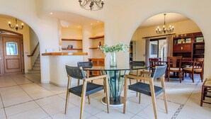 A spacious dining area with a glass-top table and four chairs, a chandelier, and a bookshelf in the background. - Resitour - Kamares Village (Tala)