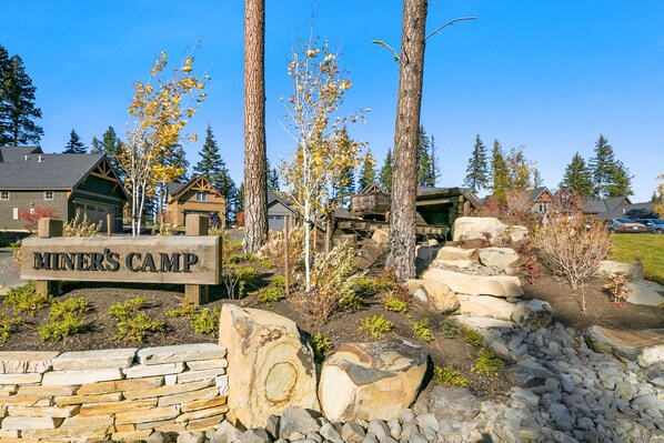 A wooden sign reading "MINER'S CAMP" with a rustic cabin and trees in the background.