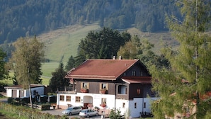 Un edificio de dos pisos con tejado de tejas rojas, rodeado de árboles y un paisaje montañoso.