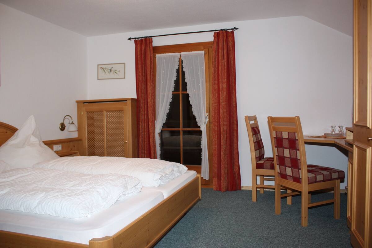 A bedroom with a wooden bed, a desk with a chair, and a window with curtains.