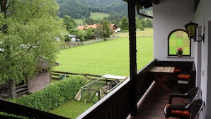 Un balcón con mesa y sillas de madera, un pequeño jardín y vistas a un campo verde y casas a lo lejos.