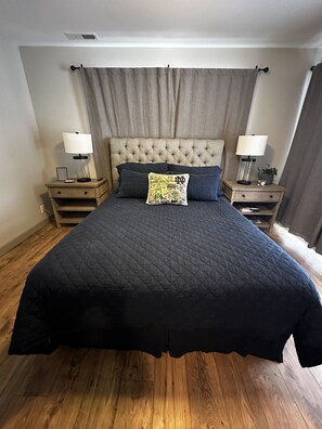 Een slaapkamer met een geborduurde hoofdbord, een donkere gewatteerde spreideken, twee nachtkastjes met lampen, en een decoratief kussen.