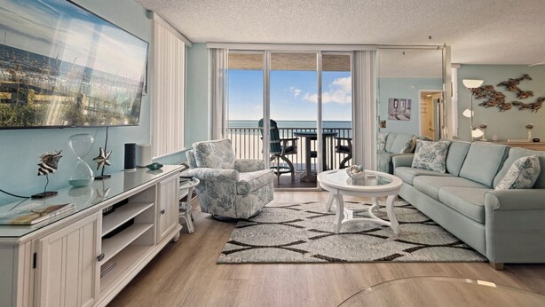 Uma sala de estar ampla com uma grande janela que oferece vista para o oceano, uma varanda com mobiliário externo e um design de interiores moderno.