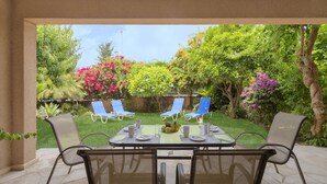 Une terrasse extérieure avec une table dressée pour deux personnes, entourée de chaises, et offrant une vue sur un jardin luxuriant, parsemé de diverses plantes et fleurs.