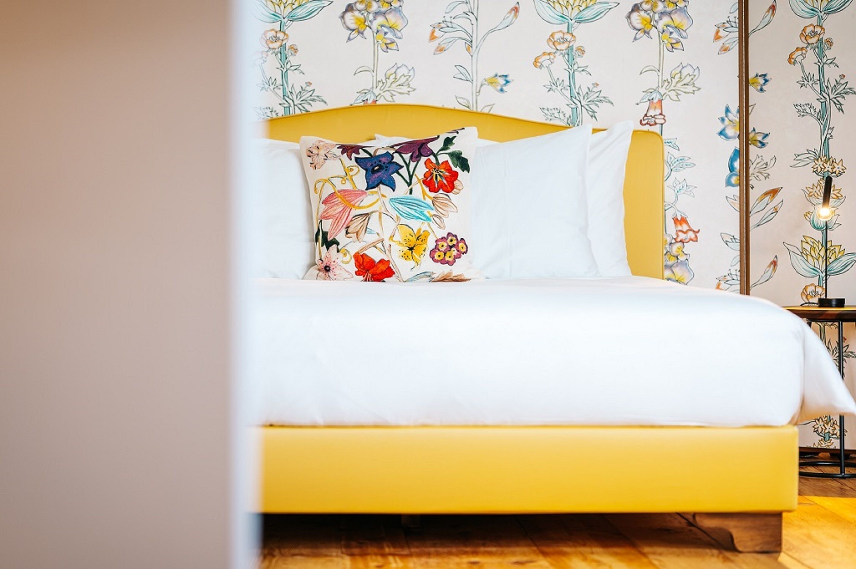 Ein Bett mit gelber Kopfteile, weißer Bettwäsche und einem blumengemusterten Kissen.