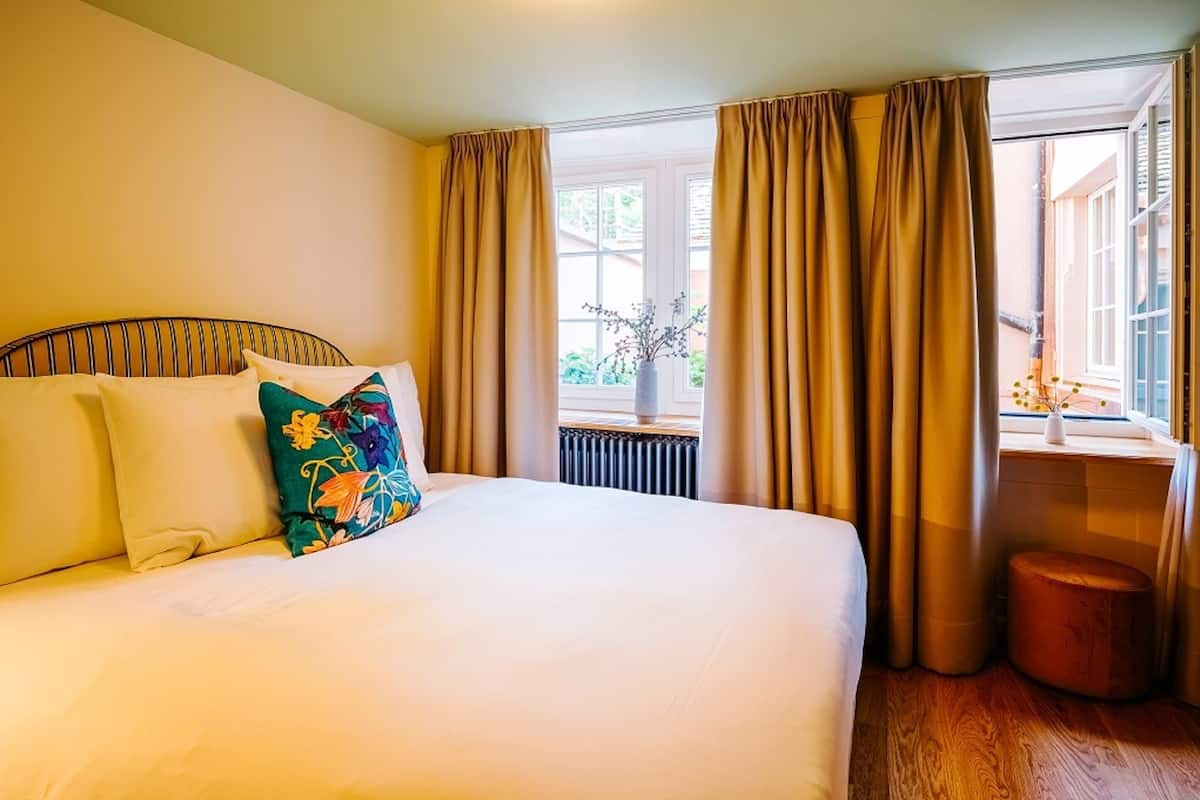 A hotel room with a large bed, beige curtains, a window with a view of greenery, and a small wooden stool.