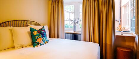 Habitación de hotel con una cama grande, cortinas color beige, una ventana con vistas a zonas verdes y un taburete de madera.