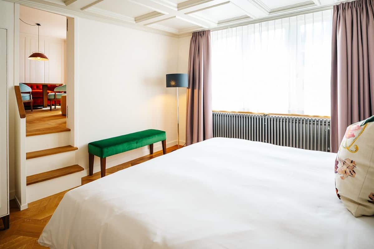 A hotel room with a large bed, a green bench, a lamp, and a view of a dining area through an open door.