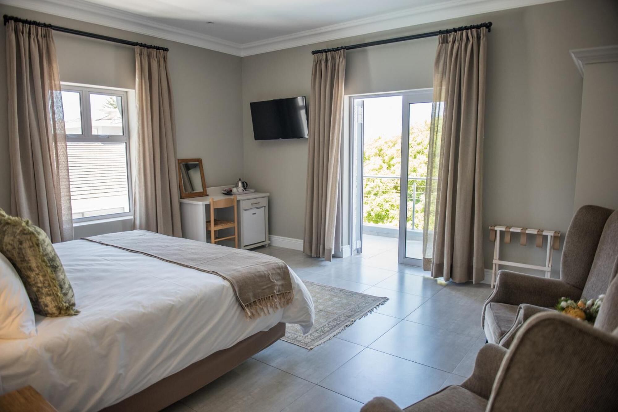 Une chambre avec un grand lit, un bureau avec une chaise, un téléviseur fixé au mur et un balcon avec des rideaux.