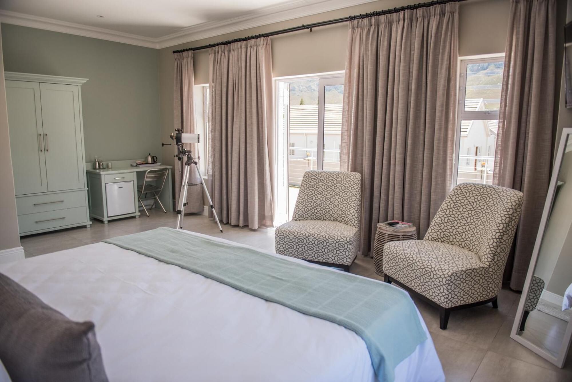 A bedroom with a bed, two patterned armchairs, a desk with a chair, a telescope, and a cabinet.
