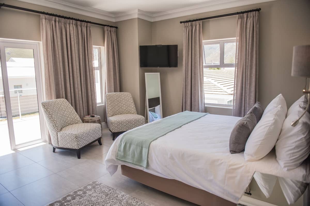 A bedroom with a bed, two patterned chairs, a flat-screen TV, and a large window with curtains.