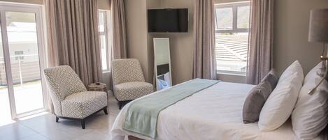 Un dormitorio con cama, dos sillas estampadas, un televisor de pantalla plana y un ventanal con cortinas.