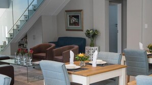 A modern dining area with a glass-top table, blue upholstered chairs, and a staircase in the background. - The Whale on Main B&B (Hermanus)
