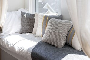 A window seat with a white bedspread, a grey knitted pillow, and a grey ribbed pillow with gold trim. - Serene, Smart, Spacious. Near Downtown + Garage (Pittsburgh)