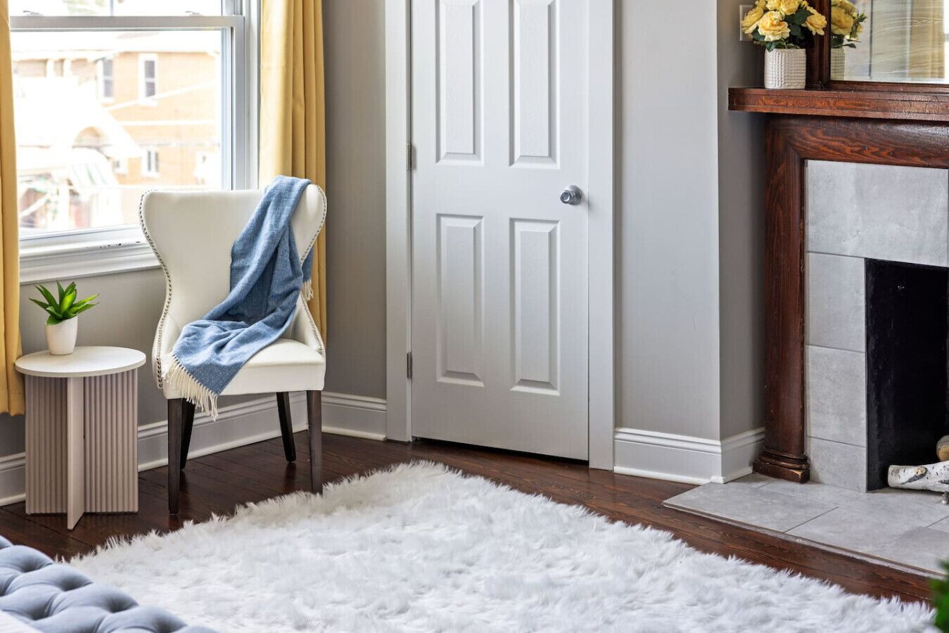 Um quarto com porta branca, tapete branco e fofo, uma cadeira com manta azul, uma pequena mesa redonda com uma planta e uma lareira com parapeito.