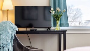 A hotel room with a flat-screen TV on a black stand, a remote control, a vase with white flowers, a blue throw blanket, and a chair. - Serene, Smart, Spacious. Near Downtown, In A Quiet Neighborhood (Pittsburgh)