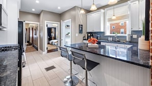A modern kitchen with a central island, bar stools, pendant lighting, and a view of a brick building outside. - Serene, Smart, Spacious. Near Downtown, In A Quiet Neighborhood (Pittsburgh)