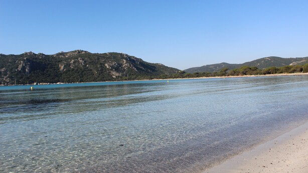 En strand med klart vatten, en sandstrand och berg i bakgrunden.