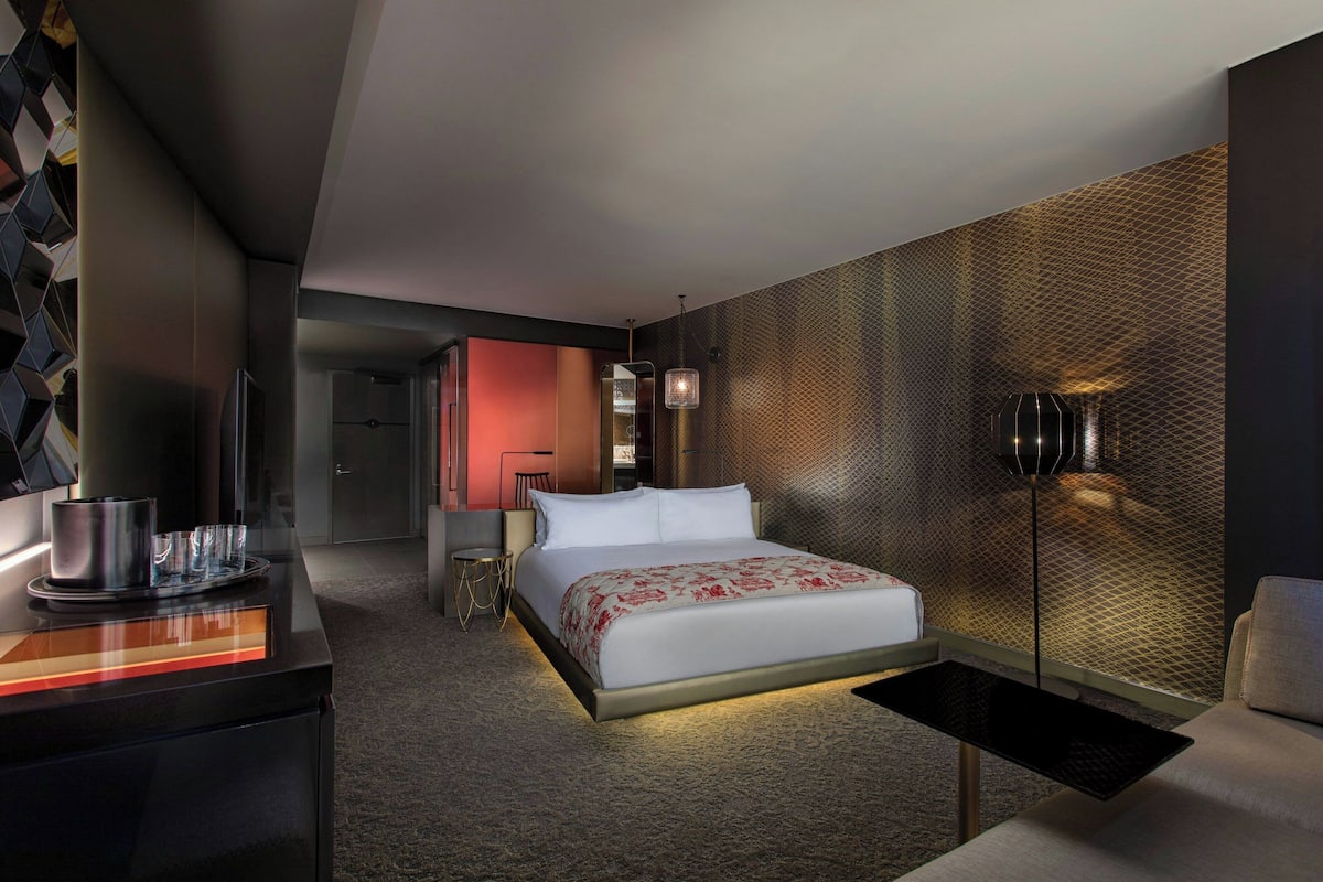 A modern hotel room with a large bed, a desk with a lamp, a chair, and a decorative wall.