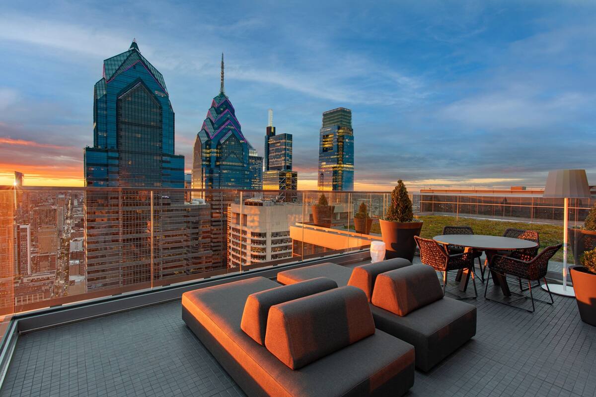 A rooftop terrace with a city skyline view, outdoor seating, and potted plants.