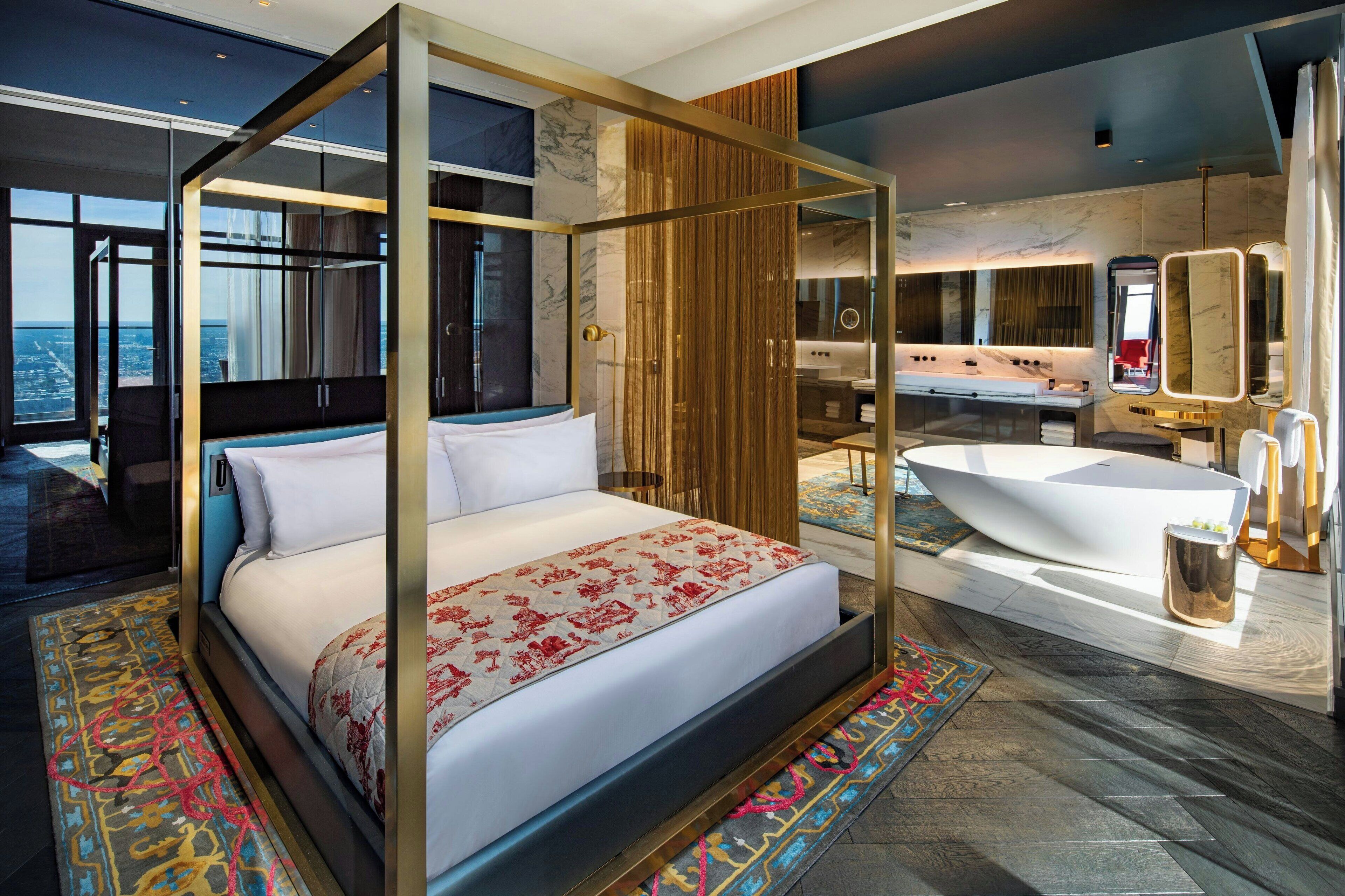 A modern hotel room with a four-poster bed, a large bathtub, and a view of the city.