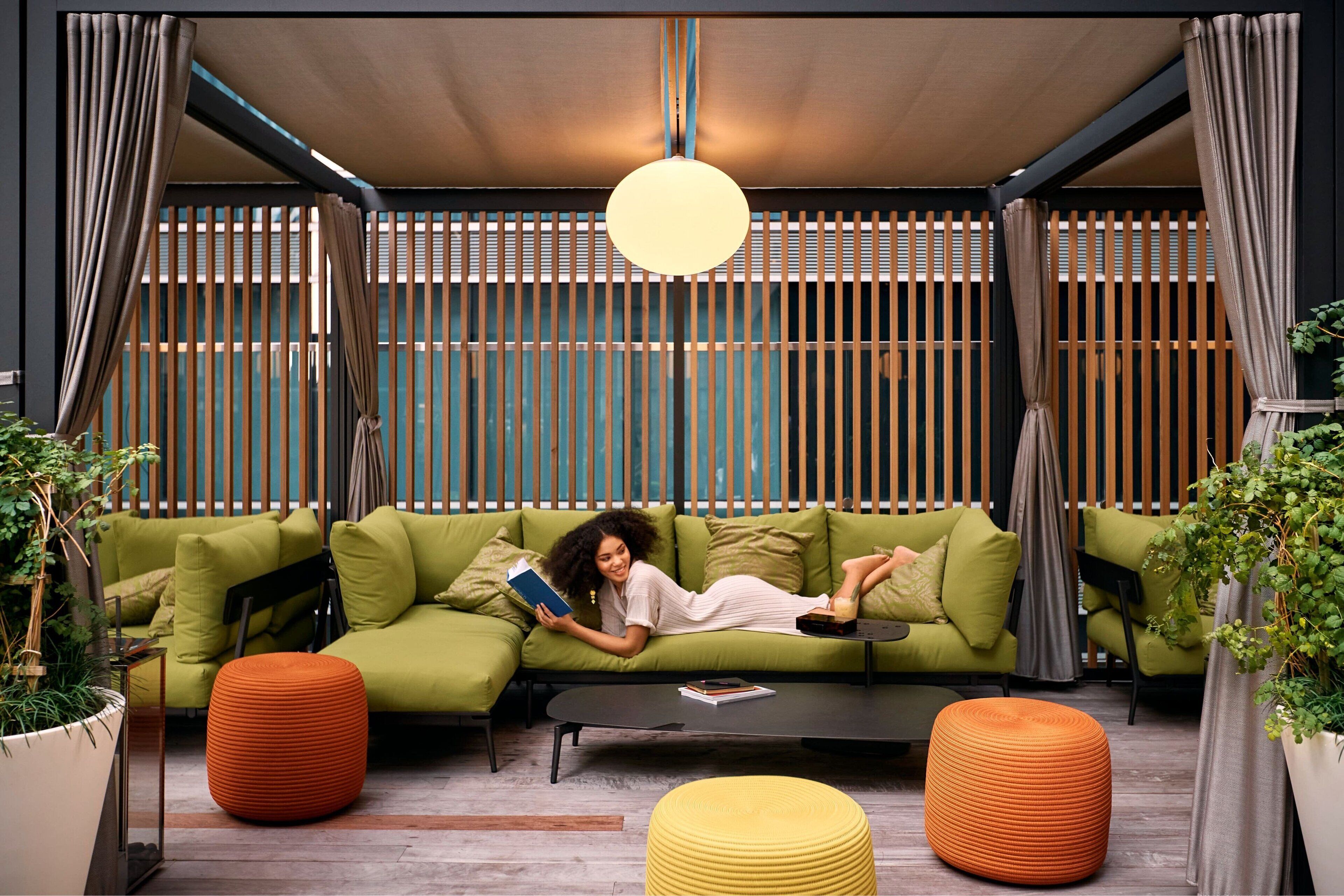 A modern living room with a green sofa, orange and yellow ottomans, and a hanging light fixture.