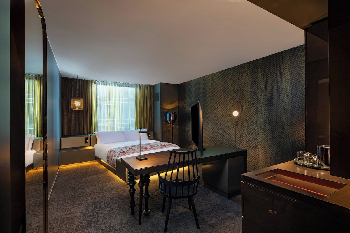 A modern hotel room with a bed, desk, chair, and a small kitchenette.