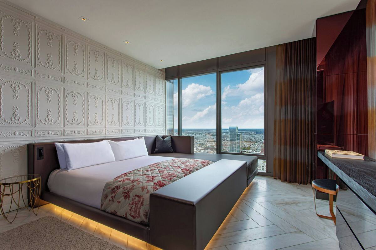 A modern bedroom with a large bed, a desk, and a city view.