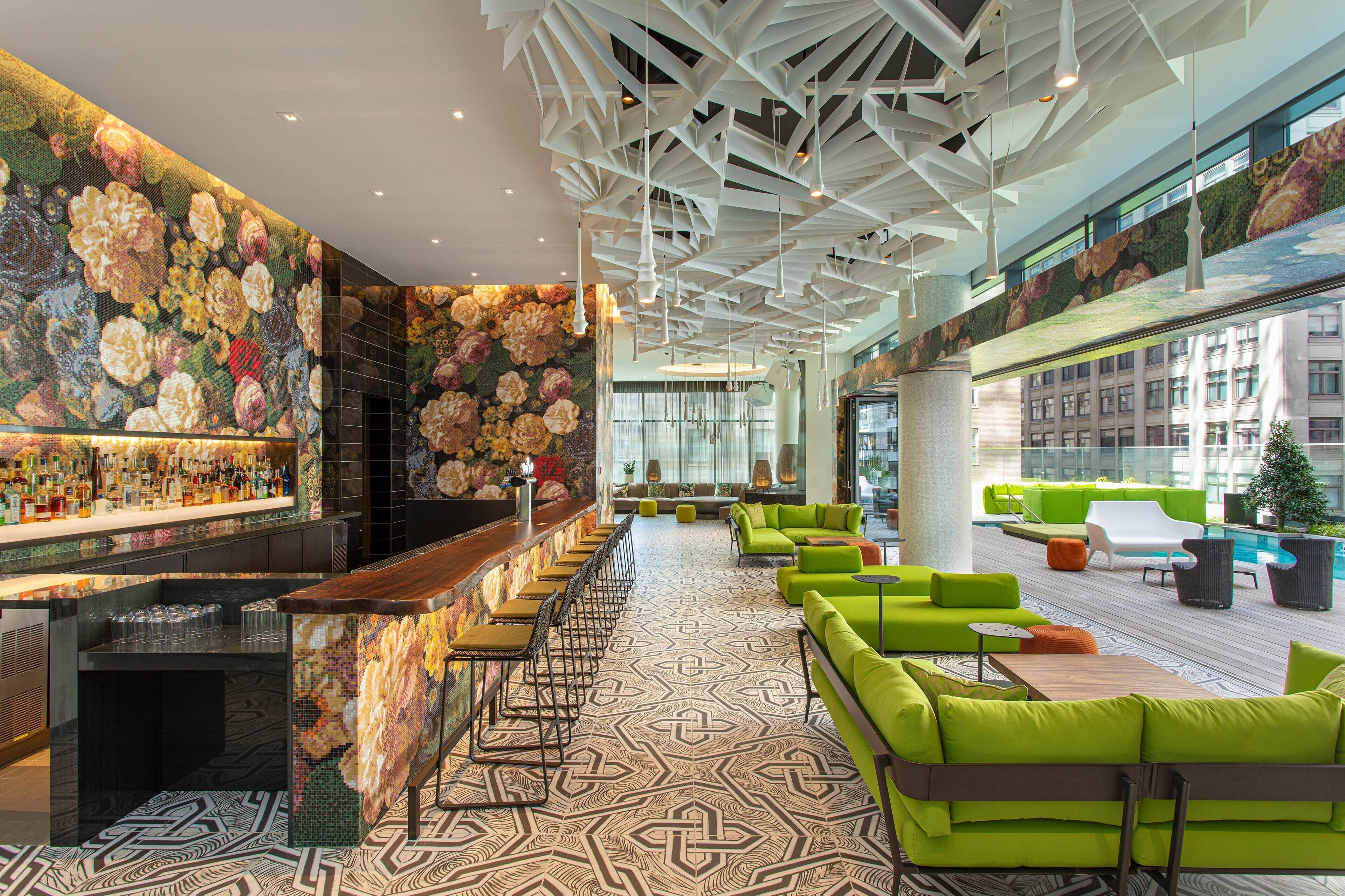 Zona de bar moderna con una barra alargada, asientos con sofás verdes y suelo estampado.