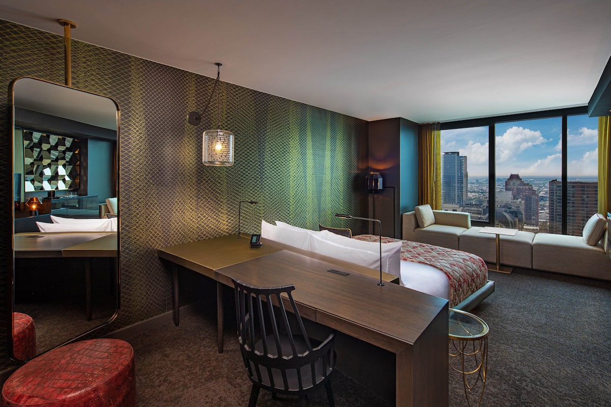 A hotel room with a bed, a desk, a chair, a small table, a mirror, and a view of the city.