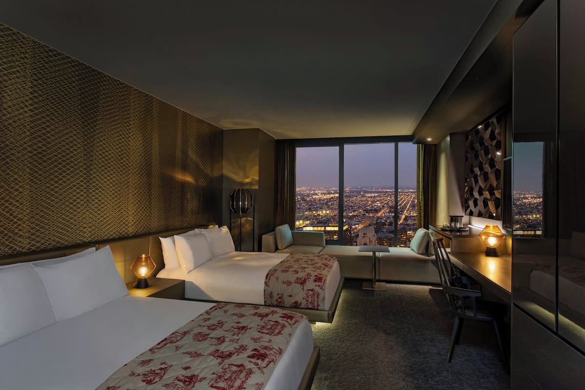 A modern hotel room with two beds, a desk, and a view of a cityscape at night.