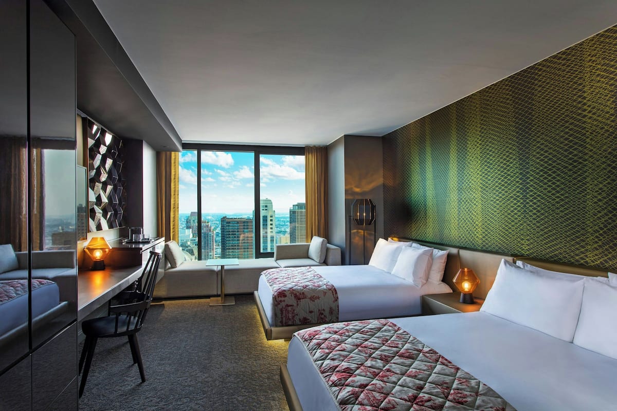 A hotel room with two beds, a desk, and a chair. There is a large window with a city view.