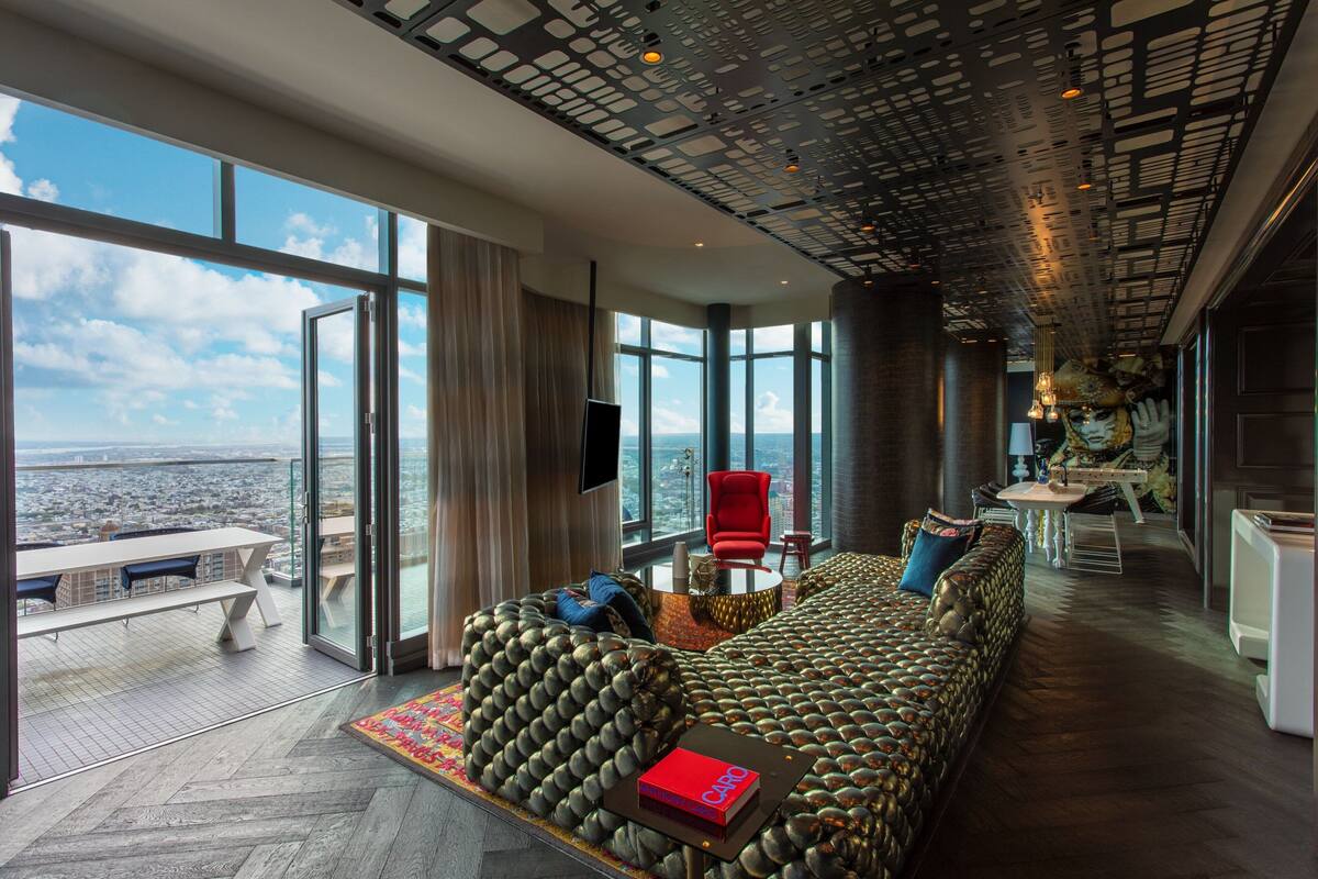 A modern hotel room with a large panoramic window, a plush sofa, a dining area, and a unique ceiling design.