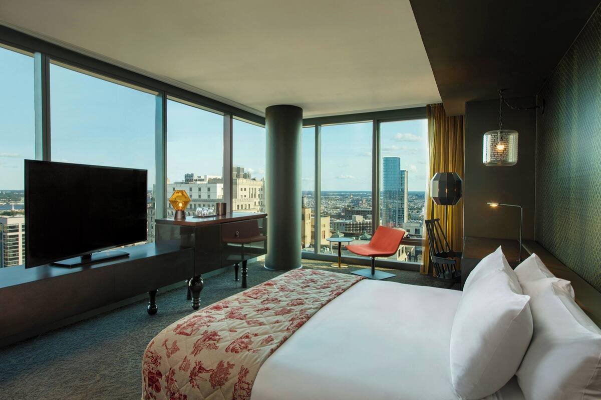 A hotel room with a large bed, a desk with a TV, a chair, and a view of the city.