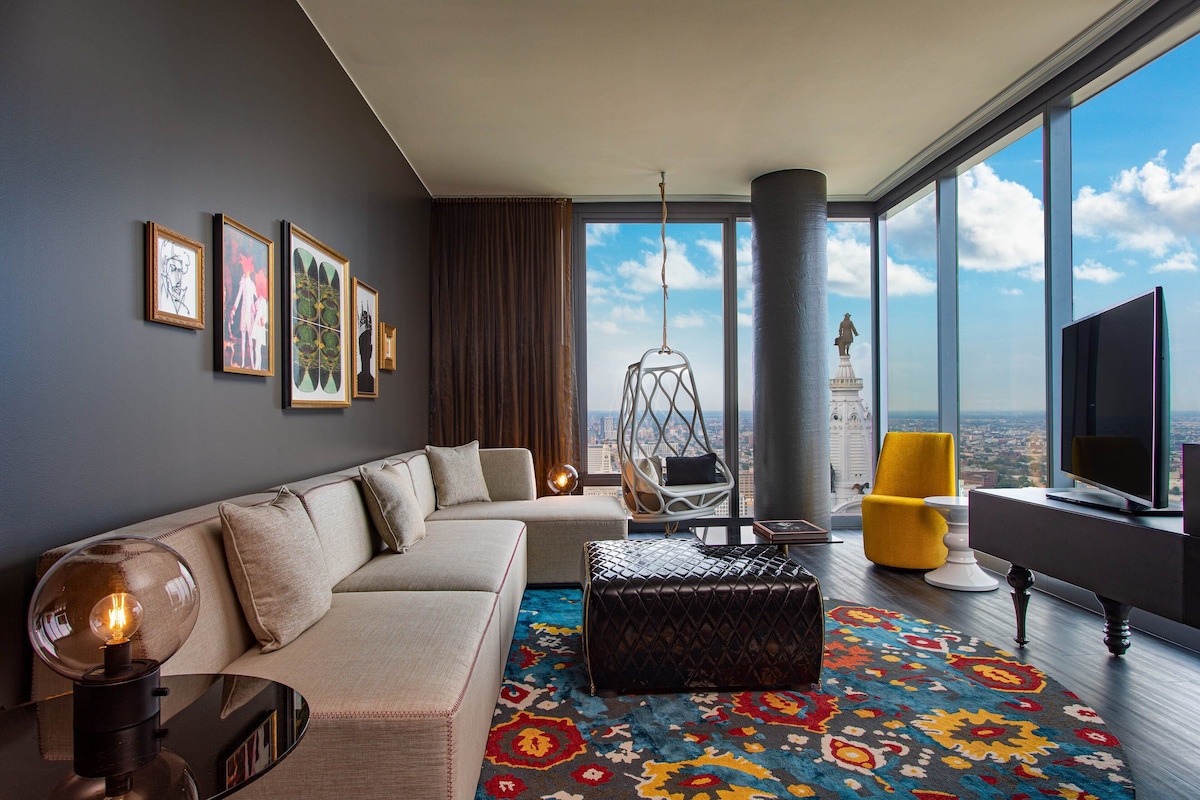 A modern living room with a sofa, ottoman, and a hanging chair. There are framed artworks on the wall, a large window with a view, and a television.
