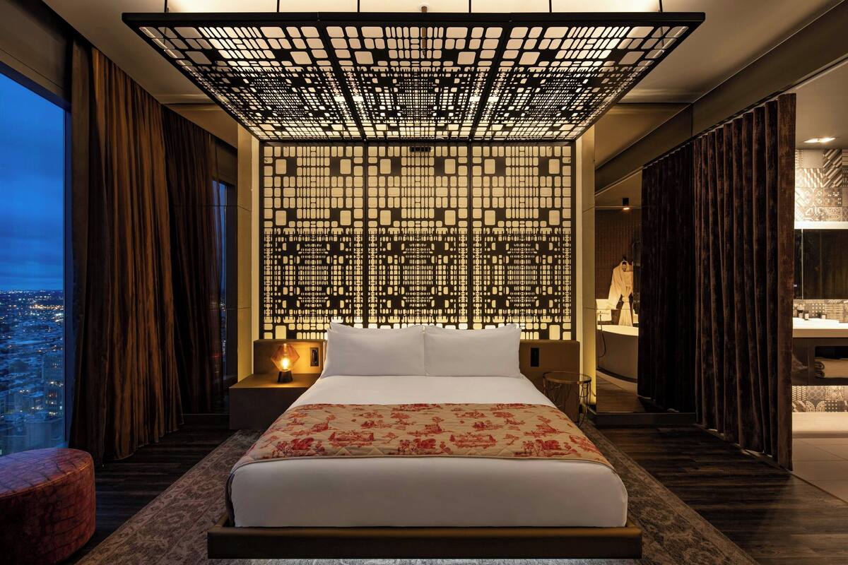 A modern hotel room with a large bed, a city view, a decorative wall, and a nightstand with a lamp.